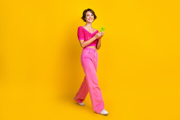 Fototapeta na wymiar Full size photo of optimistic lovely nice girl wear knit top pink pants holding smartphone walking isolated on yellow color background