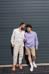 Full length of positive and young same sex couple in casual clothes holding hands while standing together near grey building on urban street