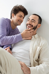 Smiling gay man with braces on teeth touching face of bearded brunette boyfriend with closed eyes...