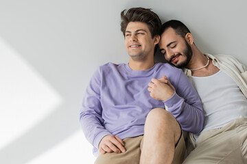 Smiling young gay man in casual clothes holding hand of bearded boyfriend with closed eyes while...