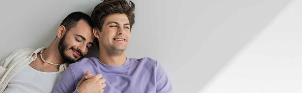 Young and carefree homosexual man with braces holding hand of bearded boyfriend and looking away while sitting together on grey background with light, banner