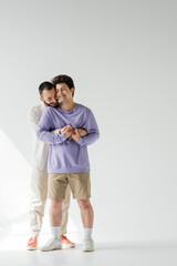 Smiling and bearded homosexual man in casual clothes holding hands of young boyfriend with closed...