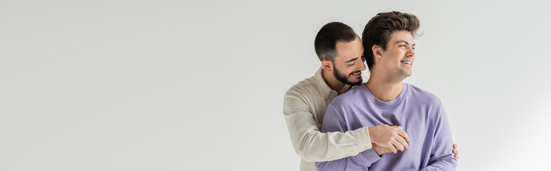 Young bearded homosexual man hugging carefree brunette boyfriend in braces and sweatshirt looking...