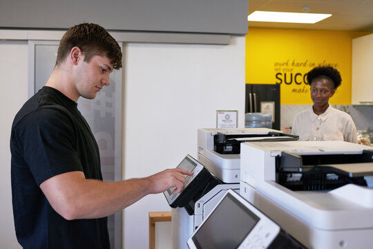 Young man uses photocopier machine in shared office workspace with colleague