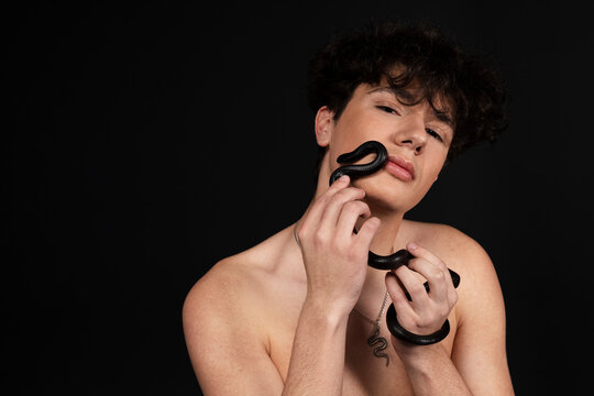 Young handsome man with naked torso, with a black snake crawling on his arms or hands. Isolated on black background.