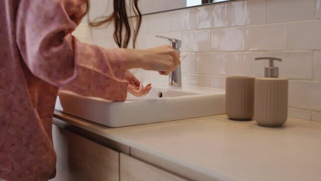 An unrecognizable woman in pajamas finishes brushing her teeth with clean water, spits into the sink and turns off the water. Morning routine. Close up, side view, horizontal 4k video