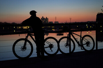 Silhouette of a cyclist standing with a bicycle near the river and admiring the sunset