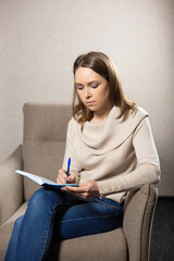 Concentrated woman makes notes in notepad sitting in comfortable chair. Psychologist writes with pen while listening to patient