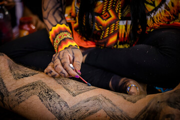 Sao Paulo, SP, Brazil - April 15 2023: Woman's hands with accessories and indigenous art doing traditional body painting of the culture with annatto and genipap details.