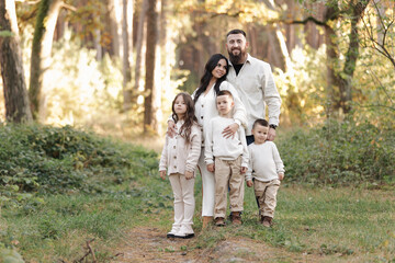 Naklejka na ściany i meble Portrait of big family outdoors. Young Stylish bearded dad with little son on shoulder, beautiful brunette mom, child boy and daughter in autumn park, smiling and looking at camera. Happy family day