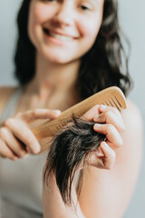 Adult young woman holding wooden hair brush and pulling healthy long black hair isolated on grey. Positive charming joy girl getting ready. Bodycare effect make hair flawless wellness shine gloss.