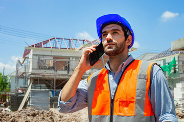 Engineer speaks on the phone. Wears a blue helmet and an orange construction vest. Engineer working on construction site with sunny background