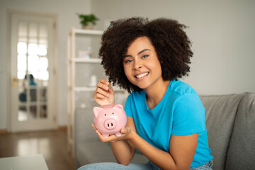 Smiling millennial curly black lady puts coins in piggy bank, for dream in living room interior
