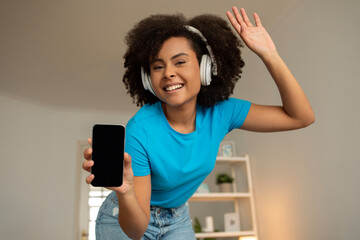 Positive young black curly woman in wireless headphones shows phone with blank screen, recommends audio app
