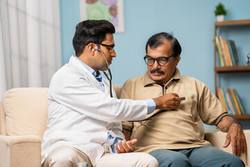 Indian doctor examining sick senior man by using stethoscope while sitting on sofa at home - concept of home medical treatment, healthcare service and routine check up.