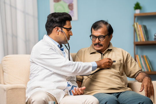 Indian doctor examining sick senior man by using stethoscope while sitting on sofa at home - concept of home medical treatment, healthcare service and routine check up.