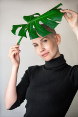 Portrait of an emotional young woman in a black turtleneck with a shaved head and a large green leaf of a flower behind which she hides. Outcome of chemotherapy in cancer treatment
