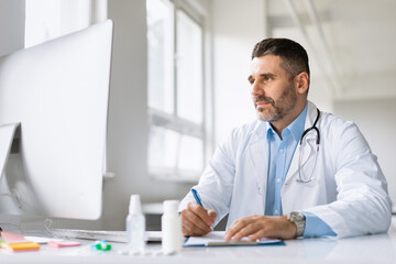 Electronic health system. Male doctor working on computer and writing prescriptions while sitting at desk in clinic