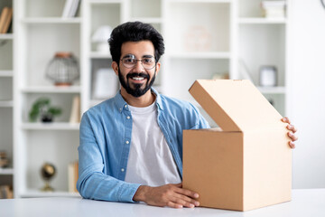 Excited indian man opening delivery box while sitting at desk at home