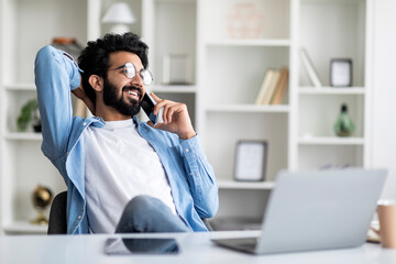 Cheerful Young Indian Guy Talking On Cellphone While Working In Home Office