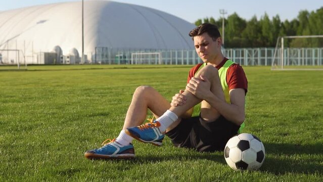 Young man football player suffering from leg injury sitting on the grass at soccer stadium.