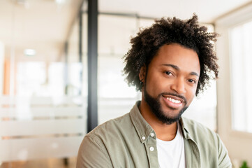 Close-up portrait of confident smiling business man standing in modern coworking office, posing and looking at the camera, curly african-american male entrepreneur, office employee