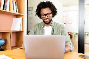 African-American guy using laptop for remote work sitting in the modern coworking space, bookshelves on the background, man in casual wear and glasses typing indoor © Vadim Pastuh