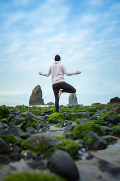 Vertical photo of woman performing yoga and meditating in tree pose on a green rocky beach in Oregon