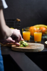 detail of a woman's hands cutting avocado in a black kitchen, orange juice in the background. girl preparing a healthy lunch.