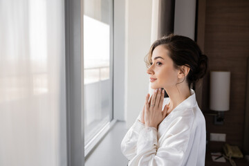 Fototapeta premium side view of hopeful young woman with engagement ring on finger standing in white silk robe with praying hands and looking through window in hotel suite, special occasion, bride on wedding day