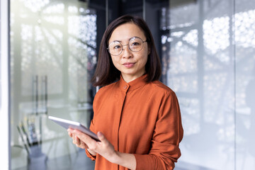 Fototapeta na wymiar Portrait of a young Asian woman programmer, developer, tester in glasses standing in the office, holding a tablet, working online and looking, posing at the camera.