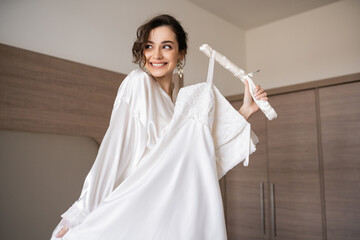 happy young bride with brunette hair in white silk robe holding soft hanger with elegant wedding dress and smiling in bedroom of hotel room, special occasion, charming woman