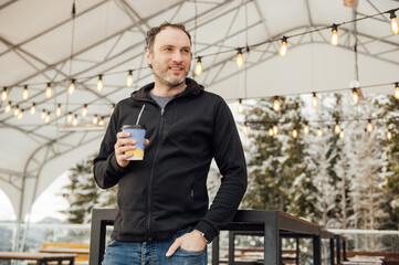 A handsome guy in a black sweater stands with a glass of coffee against the backdrop of mountains...
