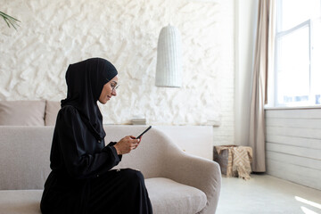 A young woman of oriental origin in a black hijab is sitting at home in a hotel and using a mobile...