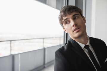 dreamy man in classic formal wear with black tie and white shirt standing in modern hotel room and looking at window, groom on wedding day, special occasion