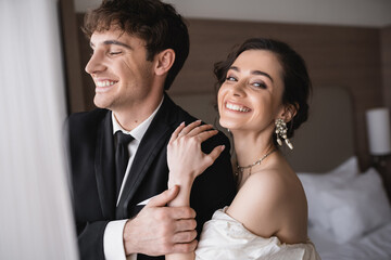 joyful bride in elegant jewelry and wedding dress hugging shoulder of happy groom in classic formal wear while standing together in modern hotel room after ceremony - Powered by Adobe