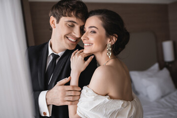 joyful young bride in jewelry and wedding dress hugging shoulder of cheerful groom in classic...
