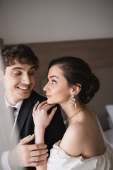 Naklejka premium pretty young bride in jewelry and wedding dress looking at cheerful groom in black formal wear while standing together in modern hotel room after ceremony