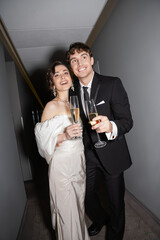 joyful groom hugging young and brunette bride in white wedding dress and holding glasses of champagne while standing and smiling together in hallway of hotel