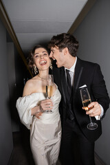 excited groom hugging young and brunette bride in white wedding dress and holding glasses of champagne while standing and smiling together in hallway of hotel, newlyweds on honeymoon © LIGHTFIELD STUDIOS
