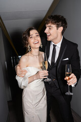 cheerful groom hugging young and brunette bride in white wedding dress and holding glasses of champagne while standing and smiling together in hallway of hotel, newlyweds on honeymoon