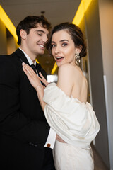happy groom in black suit hugging excited bride in white wedding dress and pearl earrings while smiling together in corridor of modern hotel, couple on honeymoon