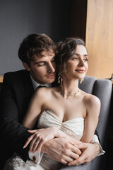 good looking groom in black suit embracing charming bride in white dress and luxurious jewelry while smiling and sitting together on comfortable armchair in hotel room