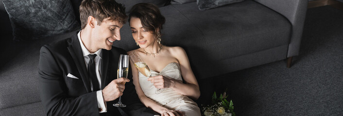 happy bride in white wedding dress and groom in black suit clinking glasses of champagne while celebrating their marriage near bridal bouquet after wedding in hotel room, banner