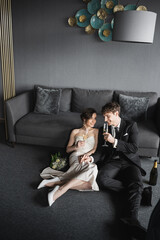 Naklejka premium cheerful bride in white dress and groom in black suit holding glasses of champagne while celebrating their marriage near bridal bouquet and couch after wedding in hotel room