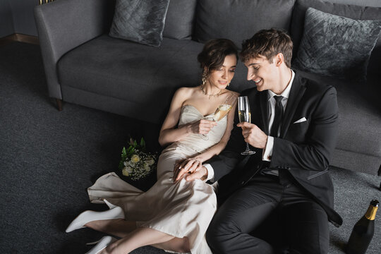 happy newlyweds in elegant attire clinking glasses of champagne while celebrating their marriage near bridal bouquet and bottle on floor after wedding in hotel room with couch