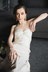 enchanting young bride with brunette hair sitting in elegant and white wedding dress, luxurious jewelry, earrings and necklace looking at camera near bridal bouquet in hotel room