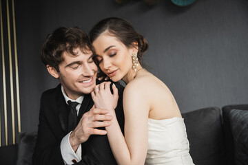 portrait of young brunette bride in luxurious earrings with pearls and white wedding dress sitting with closed eyes and hugging happy groom in black suit in hotel room