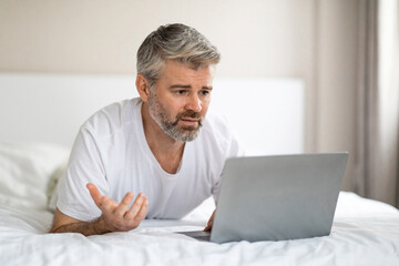 Annoyed man using laptop while relaxing on bed at home
