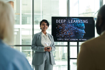Young smiling female lecturer in formalwear looking at audience while standing by interactive board...